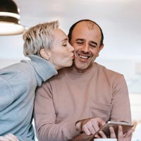 Photo couleur d'un couple assis à la table de cuisine regardant tablette, la femme embrassant l’homme - Pexels 6248562