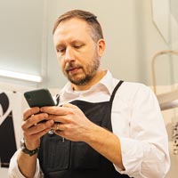 Photo couleur d'un artisan en tablier noir et chemise blanche utilisant son téléphone portable dans son atelier de travail - Pexels 6621022