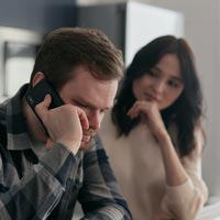 Photo en couleur d’un couple dans la cuisine avec l'homme au téléphone attablé sur une chaise haute devant un mug et des factures - Pexels 6963875