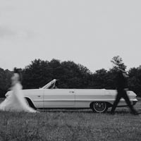 Photo en noir et blanc longue exposition d’un couple de mariés se séparant devant une voiture décapotable blanche au bord d'une route de campagne - Pexels 13627400