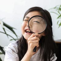 Photo en couleur d’une enfant souriante d'origine asiatique regardant d'un œil à travers une loupe dans une pièce avec des plantes vertes en arrière-plan - Pexels 5062995