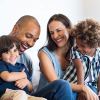Photo en couleur d’une famille de quatres personnes, deux adultes parents et deux garçons assis sur le le canapé dans leur salon en train de rire ensemble - Adobe Stock 169557997