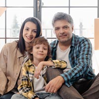 Photo en couleur d’une famille de trois personnes, deux adultes parents et leur fils assis au sol dans leur logement devant une porte vitrée, avec des cartons de déménagement derrière eux - Pexels 7414911