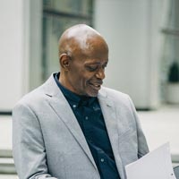 Photo en couleur d’un homme mature racisé chauve et souriant accoudé à une ballustrade de terrasse d'un immeuble de bureaux, portant une veste de costume gris clair et regardant un porte-document tenu dans ses mains - Pexels 5648396