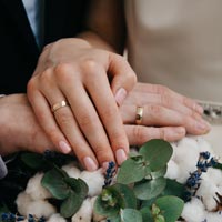 Photo en couleur de mains de mariés posés sur un bouquet de coton, avec anneaux de mariage à leur doigt - Pexels 916344